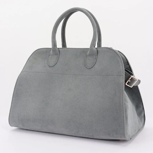May include: A gray suede handbag with two rounded handles and a structured design. The bag features a top zipper closure and a gold-colored buckle detail on the side. The bag is approximately 12 inches wide and 8 inches tall.