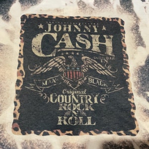 May include: Black and white graphic design with a leopard print border. The design features the text "Johnny Cash" in a vintage style font, an eagle with a shield, and the text "Original Country Rock 'n' Roll".
