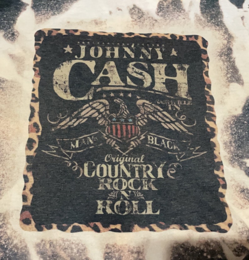 May include: Black and white graphic design with a leopard print border. The design features the text "Johnny Cash" in a vintage style font, an eagle with a shield, and the text "Original Country Rock 'n' Roll".