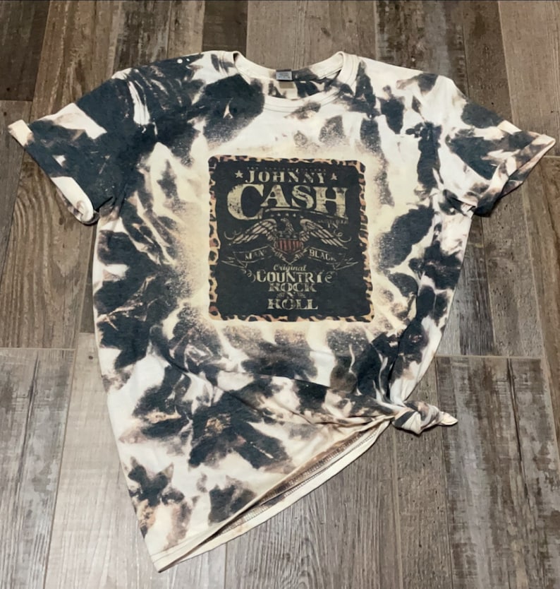 May include: A white t-shirt with a black and white tie-dye pattern. The shirt features a vintage-style graphic with the text "Johnny Cash" and "Original Countrie Rock 'n' Roll".