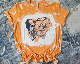 May include: Orange tie-dye t-shirt with a bleach design featuring a cartoon character, a lightning bolt, stars, and the text "Tennessee".