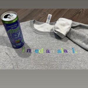 May include: A heather gray sweatshirt with the embroidered text "i need an alani." A can of Alani Nu Witch's Brew energy drink is next to the sweatshirt. The text and drink can have colorful accents.