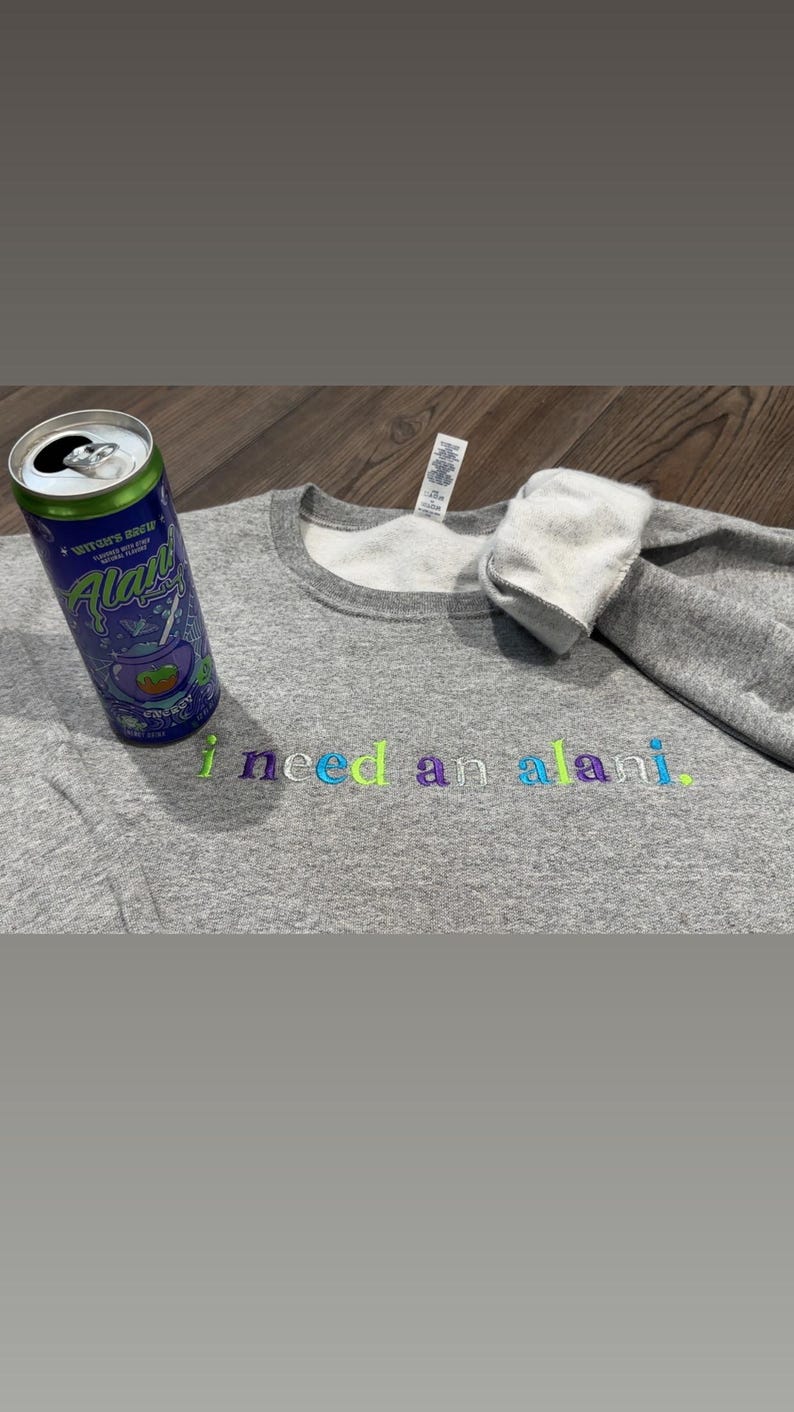 May include: A heather gray sweatshirt with the embroidered text "i need an alani." A can of Alani Nu Witch's Brew energy drink is next to the sweatshirt. The text and drink can have colorful accents.