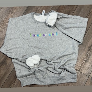 May include: A heather gray sweatshirt with the embroidered text "i need an alani" in a rainbow color scheme. The sweatshirt has a crew neck and white cuffs. The garment is laying on a wooden surface.