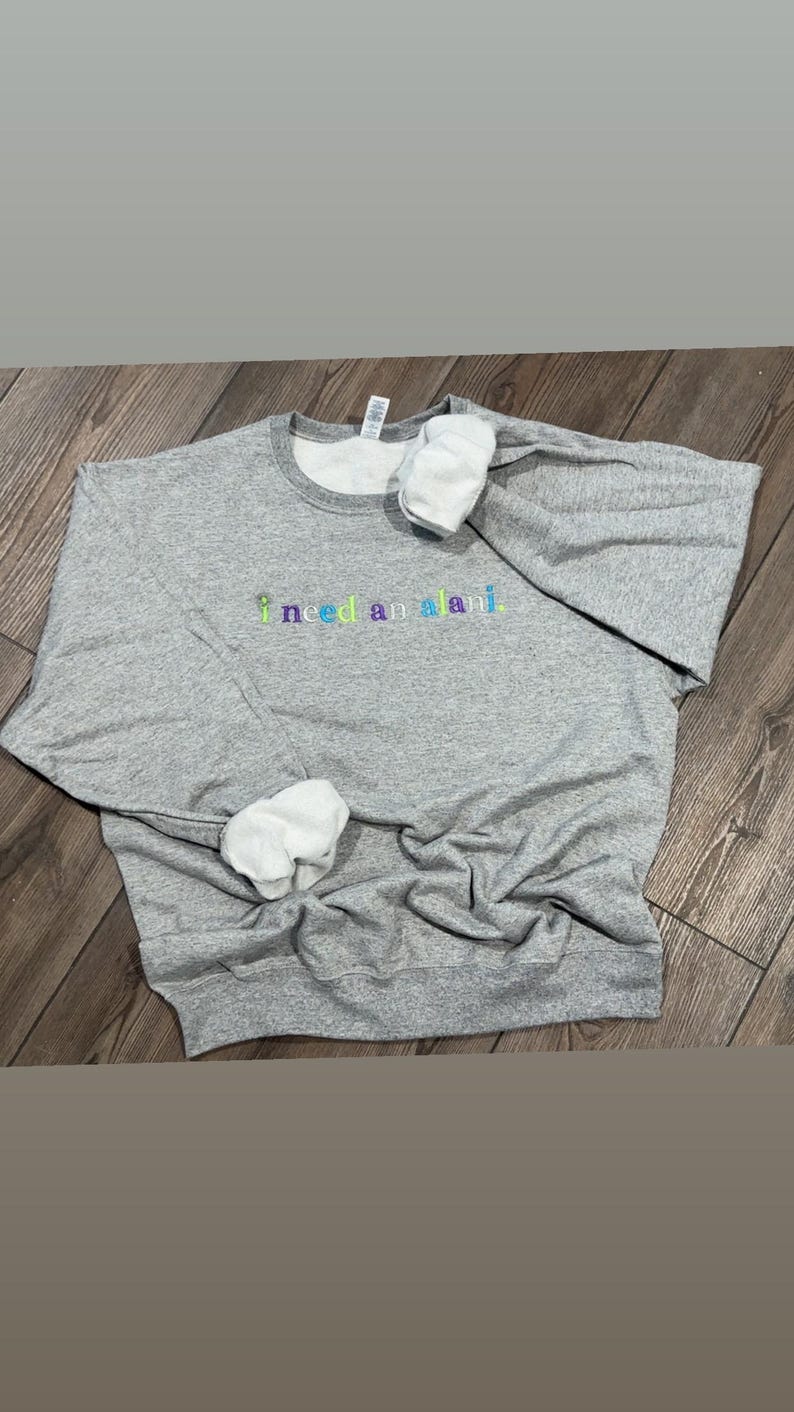 May include: A heather gray sweatshirt with the embroidered text "i need an alani" in a rainbow color scheme. The sweatshirt has a crew neck and white cuffs. The garment is laying on a wooden surface.