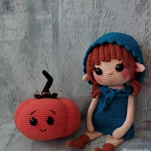 May include: A handmade crochet doll with red hair and a blue hooded dress sits beside a smiling orange pumpkin with a black stem. The doll has large black eyes and is wearing brown boots. The pumpkin and doll are on a grey background.