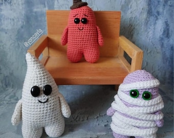 Op de afbeelding: Drie gehaakte Halloween-figuren: een geest, een mummie en een rood monster met een bruine hoed. De figuren zitten op een houten bank.