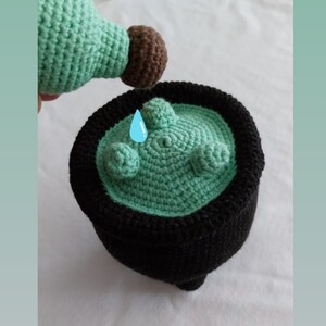 Op de afbeelding: Een groene, gehaakte ketel met een zwarte rand, die wordt bewaterd door een groene, gehaakte gieter met een bruine tuit.