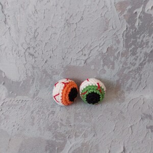 Op de afbeelding: Twee gebreide oogbollen met wit, rood, groen, oranje en zwart garen. De oogbollen liggen op een grijs, getextureerd oppervlak.
