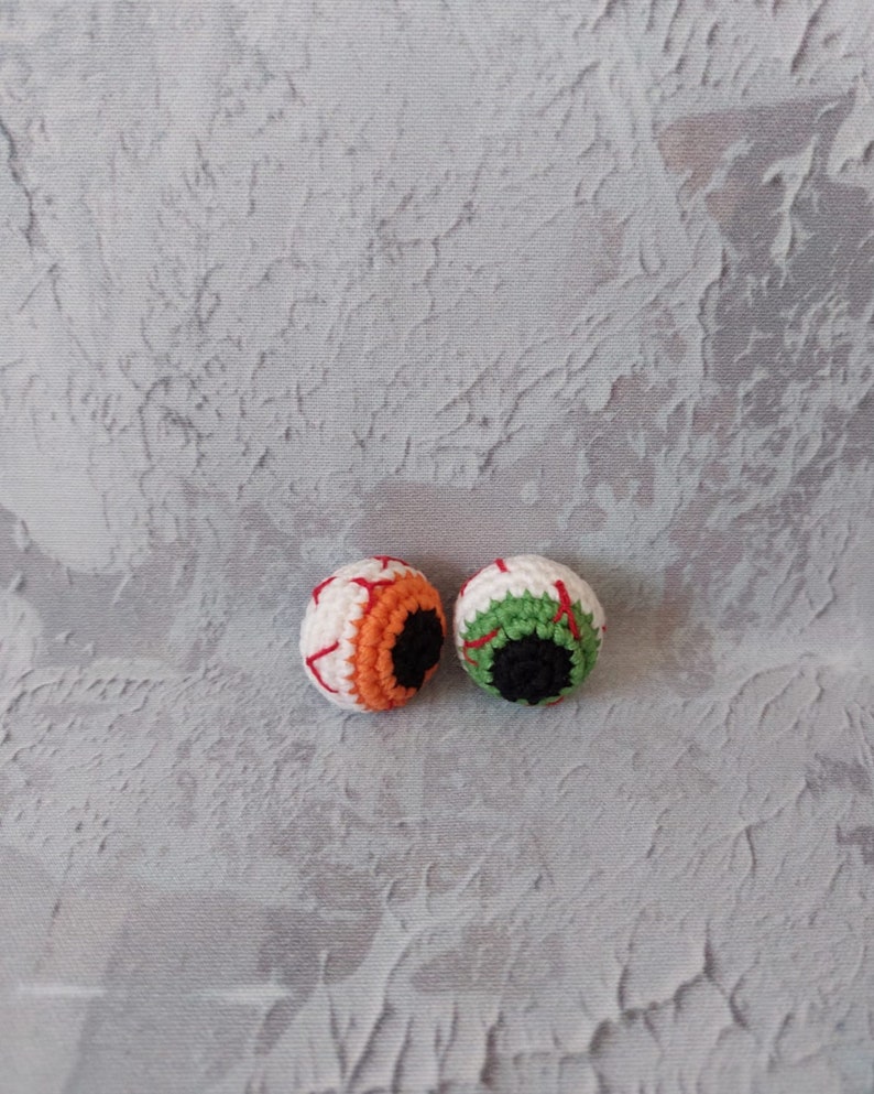 Op de afbeelding: Twee gebreide oogbollen met wit, rood, groen, oranje en zwart garen. De oogbollen liggen op een grijs, getextureerd oppervlak.