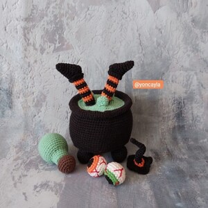 Op de afbeelding: Gecrochete heksenbenen steken uit een zwarte ketel met groene vloeistof erin. Er zijn ook gecrochete oogbollen, een groene drankfles en een zwarte heksenhoed.