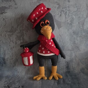 Puede incluir: Una muñeca de pájaro de ganchillo hecha a mano con un sombrero y bufanda rojos, adornada con patrones de estrellas blancas. El pájaro tiene ojos naranjas, un pico amarillo y gafas de montura metálica. Sostiene una linterna roja y blanca. La muñeca lleva un suéter rojo con estrellas blancas.