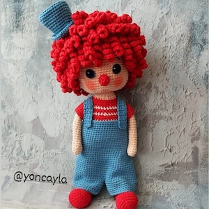May include: A crocheted clown doll with red curly hair, a blue hat, and a red and blue outfit. The doll is standing on a gray textured surface. @yoncayia