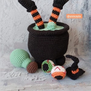 Puede incluir: Un caldero de crochet negro con líquido verde en su interior y un par de piernas de bruja que sobresalen. También hay ojos de crochet, un sombrero de bruja y una bola verde.