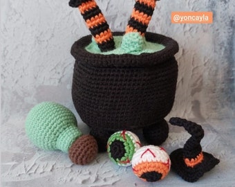 May include: A black crocheted cauldron with green liquid inside and a pair of witch legs sticking out. There are also crocheted eyeballs, a witch hat, and a green ball.