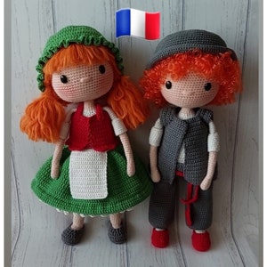 May include: Two crocheted dolls, one wearing a green dress and a red waistcoat, the other wearing a grey waistcoat and trousers. Both dolls have red hair and are standing on a wooden surface. A small French flag is in the background.