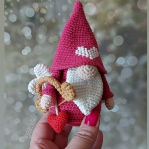 Pode incluir: Um gnomo de crochê rosa segurando uma flecha em forma de coração e um coração vermelho. O gnomo tem barba branca e um coração branco no chapéu.