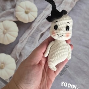 Puede incluir: Muñeca fantasma de ganchillo con sombrero de bruja negro, mejillas rosadas y ojos de botón negros. La muñeca se sostiene en una mano, con el texto "Boo! Boo!" y un emoji de fantasma. Hay calabazas blancas en el fondo.