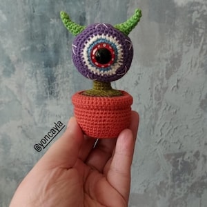 May include: A crocheted plant with a purple eye-shaped head, green horns, and a red pupil. The plant is in a small orange pot. The eye has white, blue, and red details. The plant is held in a hand.