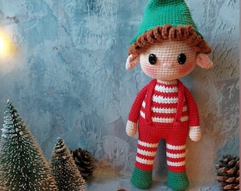 May include: A crocheted elf doll with a red and white striped outfit, green hat, and brown hair. The elf is standing on a wooden surface with pine cones and small artificial Christmas trees.