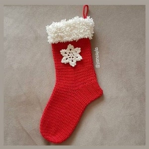 Puede incluir: Una media de Navidad roja de ganchillo con un puño blanco esponjoso y un copo de nieve blanco de ganchillo.