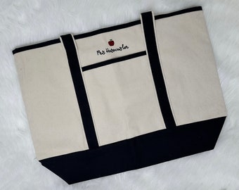 May include: A white canvas tote bag with black trim and a red apple embroidered on the front. The bag has a pocket on the front with the text "Mrs. Habeckuster" embroidered on it.