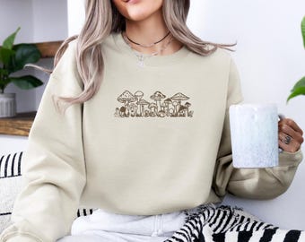 May include: A beige sweatshirt with a brown mushroom graphic. The mushrooms are arranged in a row and have different shapes and sizes. The sweatshirt has a crew neck and long sleeves.