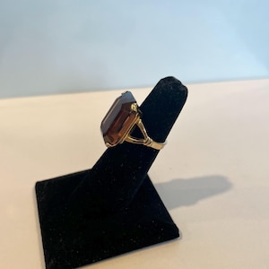 May include: A gold ring with a large, dark brown, emerald-cut gemstone. The ring is on a black ring display stand.