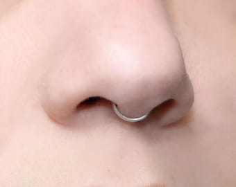 Puede incluir: Un anillo de septum circular de tono plateado usado en la nariz. La joya consiste en un aro de metal fino y liso que forma un círculo continuo, colocado de forma segura dentro del área del tabique nasal, mostrando un acabado metálico pulido.