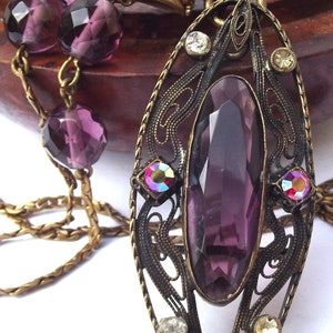 May include: A purple gemstone pendant necklace with a filigree design and a gold chain. The pendant features a large, faceted purple gemstone surrounded by smaller, iridescent gemstones.