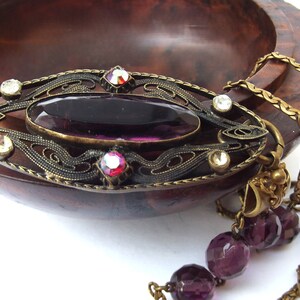 May include: A vintage-style pendant necklace with a large, oval-shaped purple glass stone set in an ornate brass frame. The frame is decorated with smaller clear and red rhinestones. The necklace is attached to a chain with a series of purple glass beads.