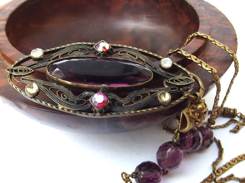 May include: A vintage-style pendant necklace with a large, oval-shaped purple glass stone set in an ornate brass frame. The frame is decorated with smaller clear and red rhinestones. The necklace is attached to a chain with a series of purple glass beads.