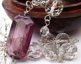 May include: A purple faceted gemstone pendant hangs from a silver chain with clear faceted crystal beads. The pendant is surrounded by clear faceted crystal beads.