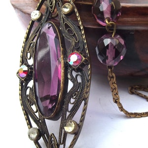 May include: A vintage pendant necklace with a large, oval-shaped purple glass stone set in an ornate, filigree frame. The pendant is attached to a gold chain with purple glass beads.
