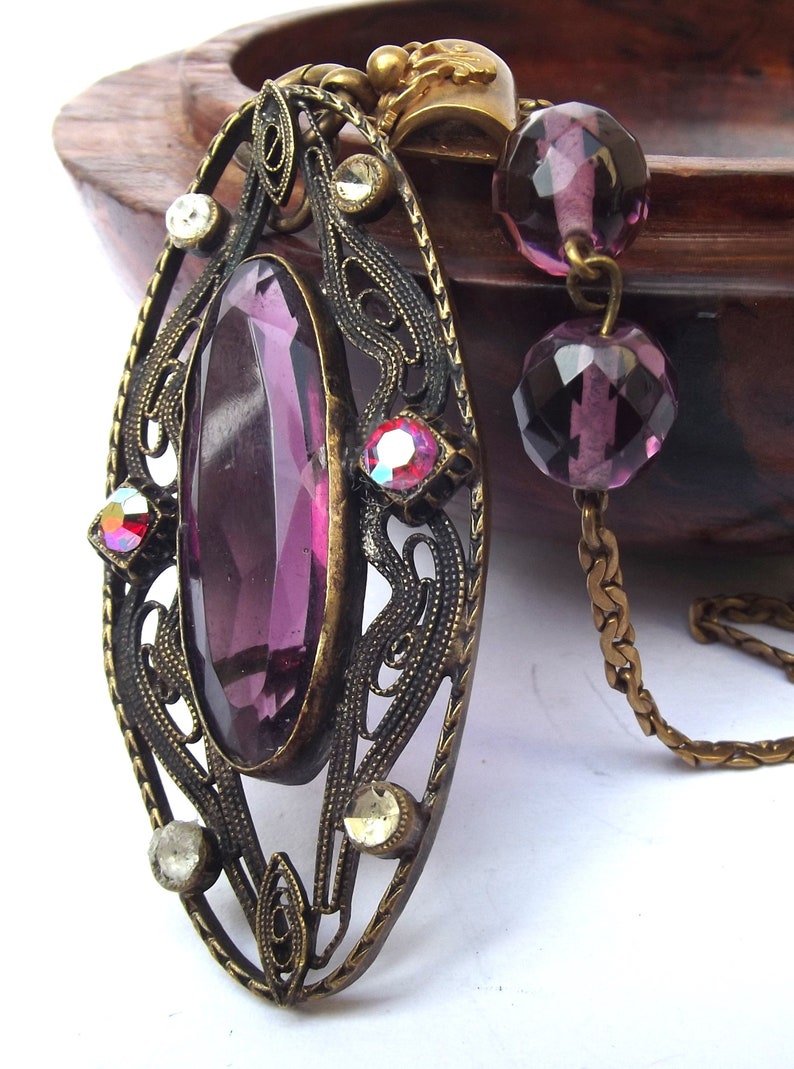 May include: A vintage pendant necklace with a large, oval-shaped purple glass stone set in an ornate, filigree frame. The pendant is attached to a gold chain with purple glass beads.