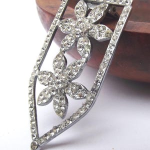 May include: A silver-toned brooch with a floral design. The brooch features two flower shapes, each composed of numerous clear, sparkling rhinestones. The brooch is set within a rectangular frame, also embellished with rhinestones, and has a pin closure on the back.