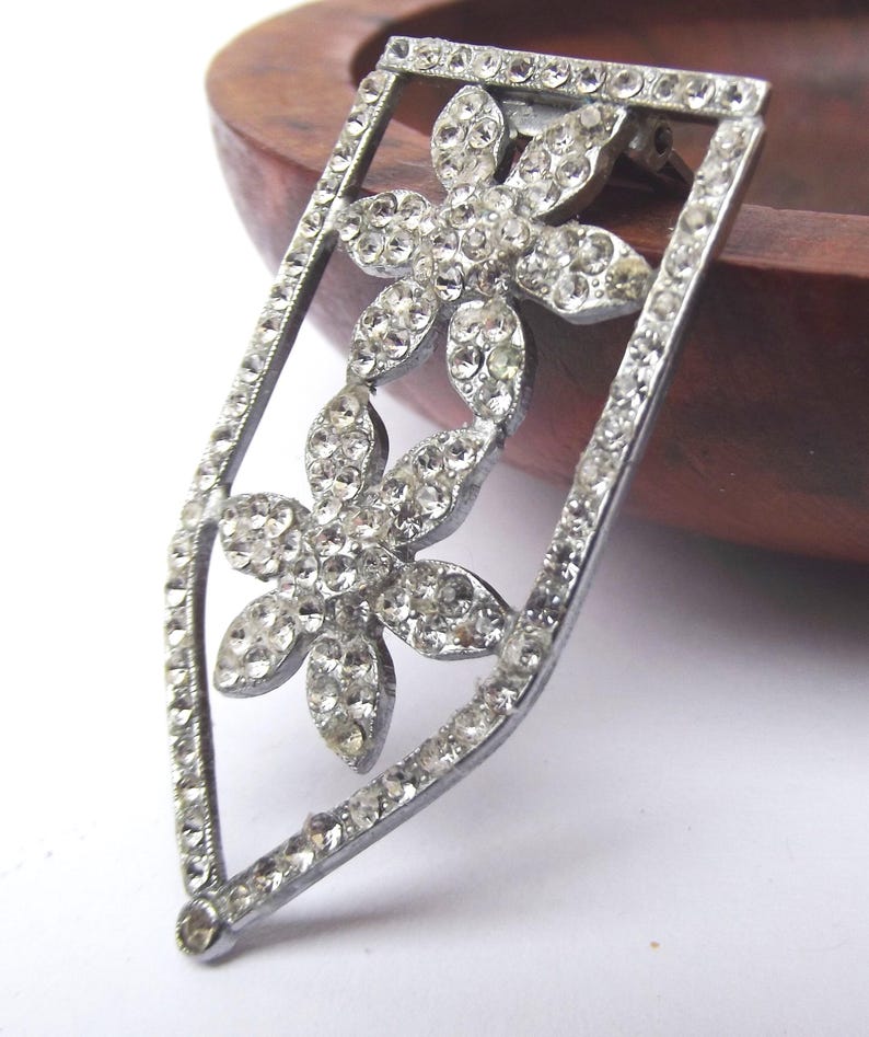 May include: A silver-toned brooch with a floral design. The brooch features two flower shapes, each composed of numerous clear, sparkling rhinestones. The brooch is set within a rectangular frame, also embellished with rhinestones, and has a pin closure on the back.