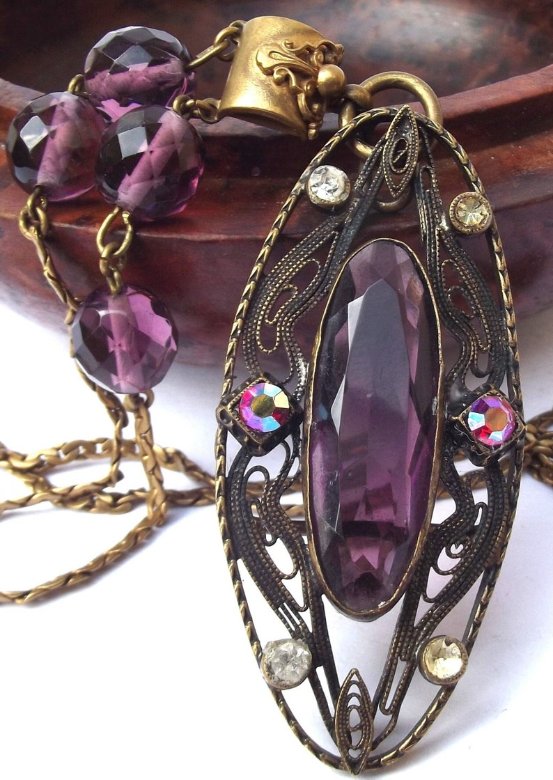 May include: A purple gemstone pendant necklace with a filigree design and a gold chain. The pendant features a large, faceted purple gemstone surrounded by smaller, iridescent gemstones.