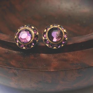 May include: A pair of round stud earrings featuring a central faceted purple stone surrounded by a ring of smaller dark purple crystals set in a circular brass-toned metal frame. The earrings have a classic design with a polished finish.