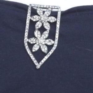 May include: A silver-toned clasp with two flower designs, each adorned with sparkling rhinestones. The clasp is set against a dark blue fabric, creating a striking contrast. The clasp is likely for a handbag or other accessory.