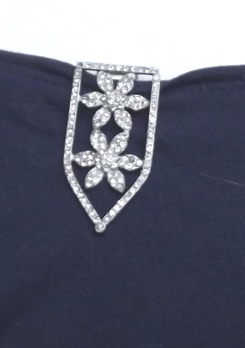 May include: A silver-toned clasp with two flower designs, each adorned with sparkling rhinestones. The clasp is set against a dark blue fabric, creating a striking contrast. The clasp is likely for a handbag or other accessory.