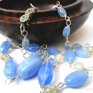 May include: A necklace featuring oval-shaped light blue glass beads interspersed with smaller, round, translucent beads containing colorful floral patterns. The beads are connected by silver-toned metal links, and the clasp is an infinity-shaped silver-toned metal piece encrusted with small clear crystals.