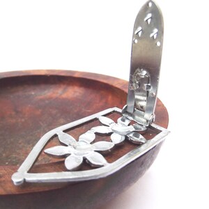 May include: A silver-toned metal clip with a floral design, featuring a rectangular frame and three flower shapes. The clip is attached to a wooden bowl with a dark brown finish. The clip is open.