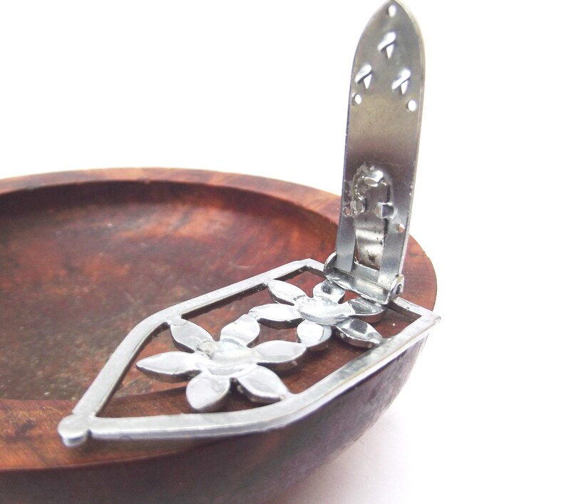 May include: A silver-toned metal clip with a floral design, featuring a rectangular frame and three flower shapes. The clip is attached to a wooden bowl with a dark brown finish. The clip is open.