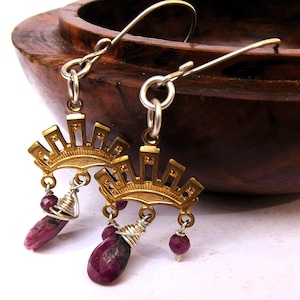 May include: Gold-coloured metal earrings with a sunburst design. Each earring has a dangling, teardrop-shaped, dark pink gemstone and smaller, round gemstones. The earrings are displayed against a wooden bowl.