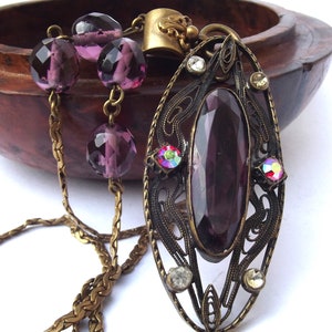 May include: A purple glass pendant necklace with a filigree design and a chain made of brass. The pendant features a large, faceted purple glass stone surrounded by smaller clear and iridescent stones.