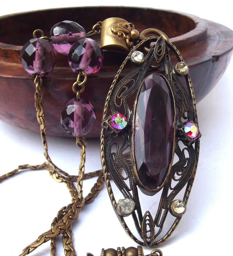 May include: A purple glass pendant necklace with a filigree design and a chain made of brass. The pendant features a large, faceted purple glass stone surrounded by smaller clear and iridescent stones.
