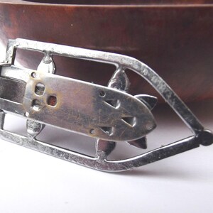 May include: A vintage silver-toned metal belt buckle with an Art Deco design. The buckle features a rectangular center with decorative cutouts and a stylized floral or geometric shape. The buckle is set against a blurred background.