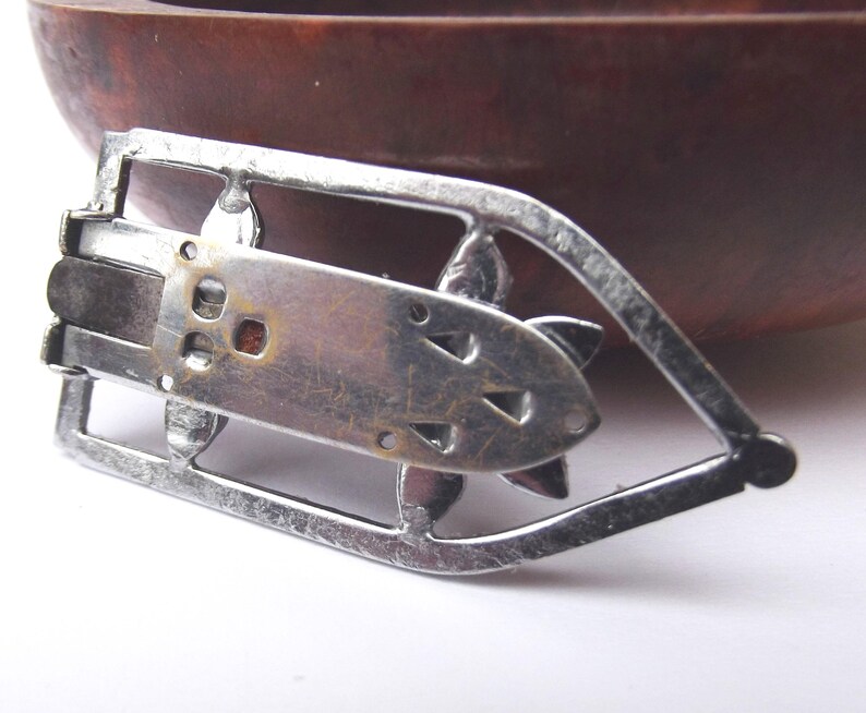 May include: A vintage silver-toned metal belt buckle with an Art Deco design. The buckle features a rectangular center with decorative cutouts and a stylized floral or geometric shape. The buckle is set against a blurred background.