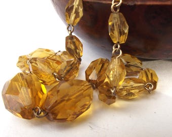 May include: Close-up of a necklace with large, faceted amber-colored beads. The beads are strung together with small gold-colored metal links. The necklace is resting on a white surface, with a dark wooden bowl in the background.